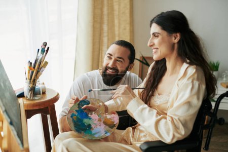 Téléchargez les photos : Mari positif aidant sa belle femme inclusive en fauteuil roulant à peindre sur chevalet pendant qu'il est à la maison - en image libre de droit