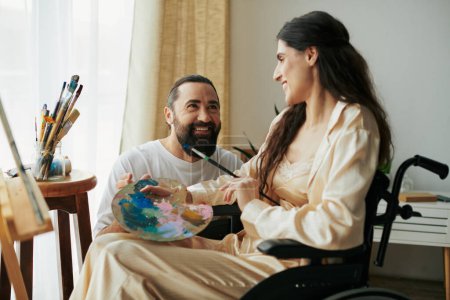 positiver Ehemann hilft seiner inklusiven schönen Frau im Rollstuhl, zu Hause auf Staffelei zu malen