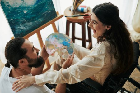 appealing woman with mobility disability painting on easel next to her cheerful bearded husband