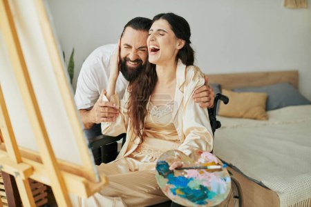 Téléchargez les photos : Attrayant couple joyeux de barbu homme et femme handicapée peinture sur chevalet ensemble à la maison - en image libre de droit