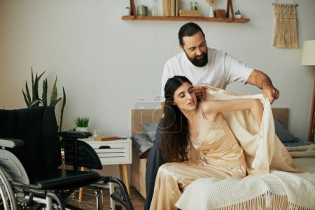 schöner liebevoller Mann in Hauskleidung, der seiner behinderten Frau hilft, sich zu Hause im Schlafzimmer in Pyjama zu verwandeln