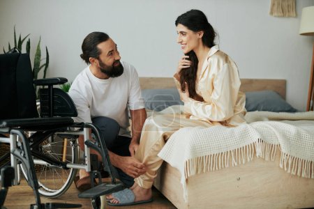 bärtiger liebevoller Mann in Hauskleidung, der seiner behinderten Frau hilft, sich zu Hause im Schlafzimmer in Pyjama zu verwandeln