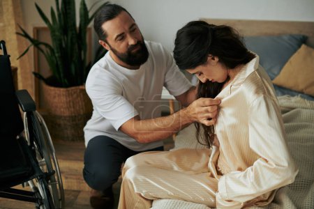 bärtiger liebevoller Mann in Hauskleidung, der seiner behinderten Frau hilft, sich zu Hause im Schlafzimmer in Pyjama zu verwandeln