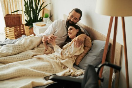 Foto de Atractiva mujer alegre con discapacidad de movilidad tumbada en la cama junto a su marido barbudo amoroso - Imagen libre de derechos