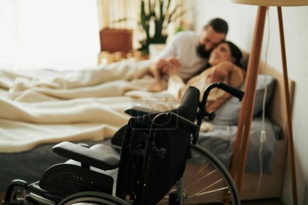 Foto de Se centran en la silla de ruedas moderna en frente de una pareja borrosa y amorosa tumbados en la cama juntos mientras están en casa - Imagen libre de derechos