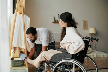 bearded loving man helping his inclusive wife on wheelchair to get dressed while in bedroom