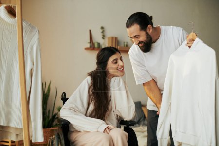 bearded caring man helping his inclusive wife on wheelchair to get dressed while in bedroom
