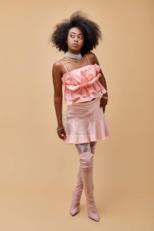verträumtes afrikanisch-amerikanisches Mädchen in den 20er Jahren posiert in pfirsichfarbenem Rüschenoberteil und Overknee-Stiefeln auf beigem Hintergrund