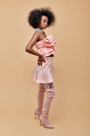 pretty african american girl in 20s posing in peach ruffle top and over-knee boots on beige backdrop