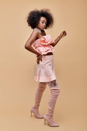 young african american girl in 20s posing in peach ruffle top and over-knee boots on beige backdrop