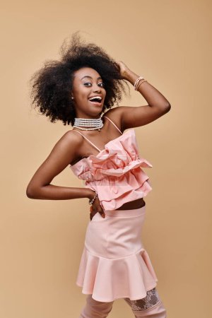 excited african american girl in 20s posing in peach ruffle top and pastel skirt with hand on hip