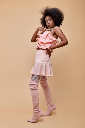 junges afrikanisch-amerikanisches Model posiert in pfirsichfarbenem Rüschenoberteil und Overknee-Stiefeln auf beigem Hintergrund