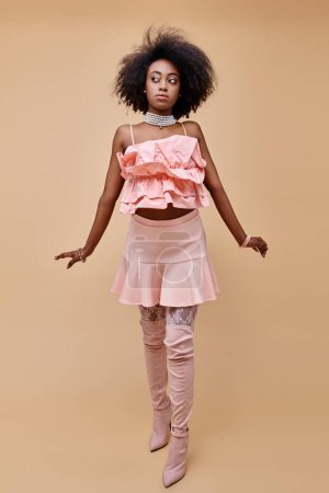 attraktives junges afrikanisch-amerikanisches Model posiert in pfirsichfarbenem Rüschenoberteil und Overknee-Stiefeln auf beige