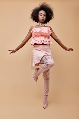 african american woman in 20s, posing in peach ruffle top and over-knee boots on beige, on one leg