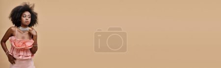 Foto de Estandarte de la mujer de piel oscura con el pelo rizado posando en melocotón pastel con volantes superior sobre fondo beige - Imagen libre de derechos