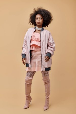 african american girl in 20s, posing in peach outfit with jacket and over-knee boots on beige, style