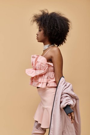 curly african american woman posing in pastel outfit with jacket on beige background, peach fuzz