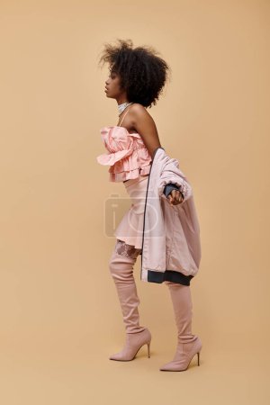 african american woman in 20s, posing in peach outfit with jacket and over-knee boots on beige