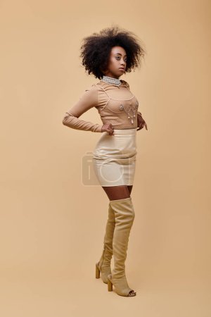 Modisches afrikanisch-amerikanisches Mädchen in pastellfarbenem Minirock und schenkelhohen Stiefeln vor beigem Hintergrund