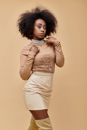 trendige afrikanisch-amerikanische Frau mit lockigem Haar posiert in stylischem Pastell-Outfit vor beigem Hintergrund