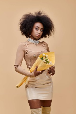 Photo for Young african american model posing in pastel peach outfit and holding flowers on beige backdrop - Royalty Free Image