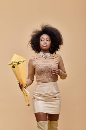 young african american model posing in pastel peach outfit and holding bouquet on beige backdrop