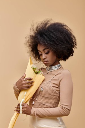 Photo for Pretty african american model posing in pastel peach outfit and holding flowers on beige backdrop - Royalty Free Image