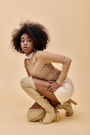 lockiges afrikanisch-amerikanisches Model in stylischem pastellfarbenem Outfit und schenkelhohen Stiefeln auf beige sitzend
