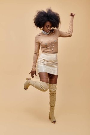 afrikanisch-amerikanisches Model im trendigen Outfit und schenkelhohen Stiefeln auf einem Bein stehend vor beigem Hintergrund