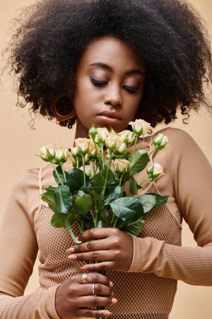 junge und lockige afrikanisch-amerikanische Frau schaut auf winzige Rosen auf beigem Hintergrund, pfirsichfarbene Fuzz