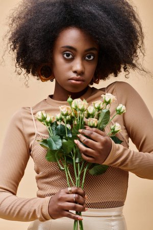 joven rizado africano americano mujer sosteniendo diminutas rosas sobre fondo beige, color de pelusa melocotón