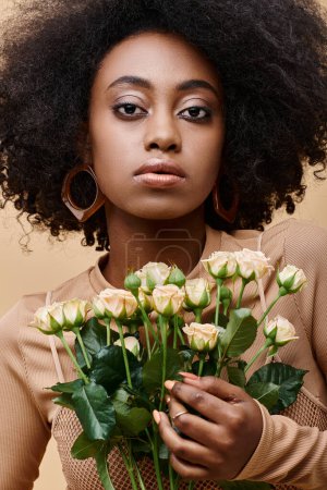 young and curly african american girl in 20s holding tiny roses on beige background, peach fuzz
