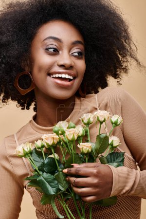 happy and curly african american woman holding tiny roses on beige background, peach fuzz color