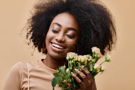 joyful curly african american woman holding tiny roses on beige background, peach fuzz color