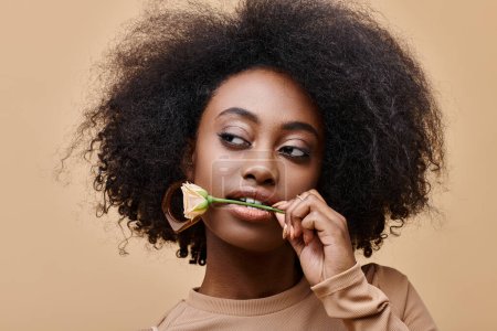 young and curly african american woman with small rose in teeth on beige background, peach fuzz