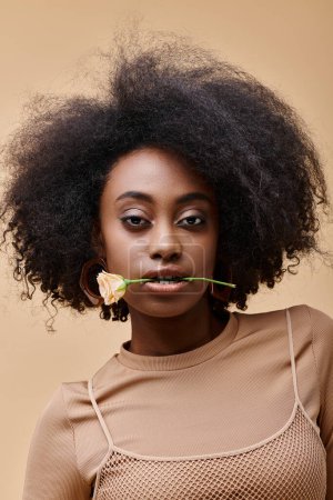 young curly african american woman with small rose in teeth on beige background, peach fuzz