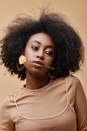daring curly african american woman with small rose in teeth on beige background, peach fuzz