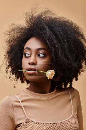 daring curly african american girl in 20s biting small rose on beige background, peach fuzz color