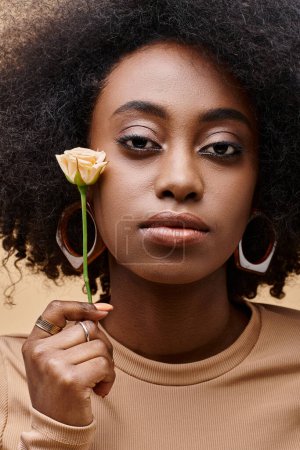 sensual and curly african american woman in 20s holding small rose on beige background, peach fuzz