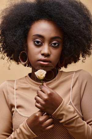 Foto de Sensual joven afroamericana chica de 20 años sosteniendo pequeña rosa sobre fondo beige, pelusa melocotón - Imagen libre de derechos