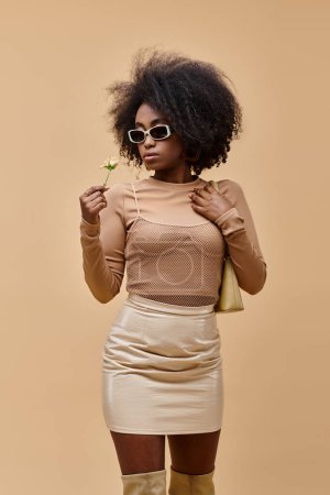 curly african american woman in sunglasses holding handbag and tiny rose on beige background