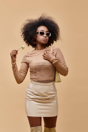 stylish african american woman in sunglasses holding handbag and tiny rose on beige background