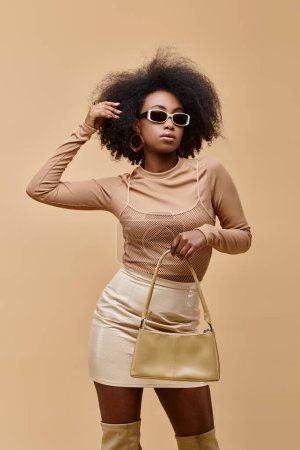 Foto de Modelo afroamericano joven y elegante en gafas de sol con bolso de moda sobre fondo beige - Imagen libre de derechos