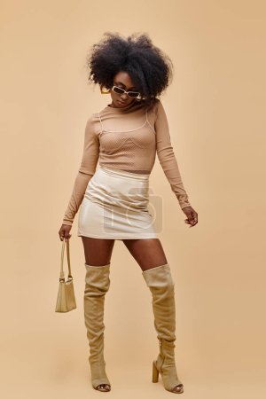 afrikanisch-amerikanisches Model in Sonnenbrille und schenkelhohen Stiefeln mit trendiger Handtasche auf beigem Hintergrund