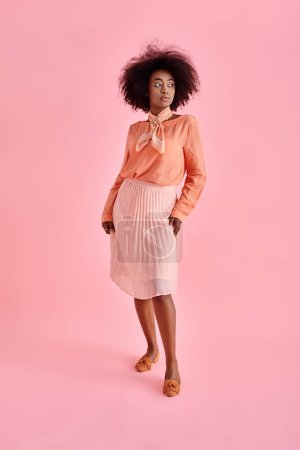elegant african american woman in peach blouse and midi skirt posing on pastel pink background