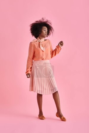 dreamy african american woman in peach blouse and midi skirt posing on pastel pink background