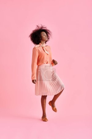 curly african american woman in peach blouse and midi skirt posing on pastel pink background