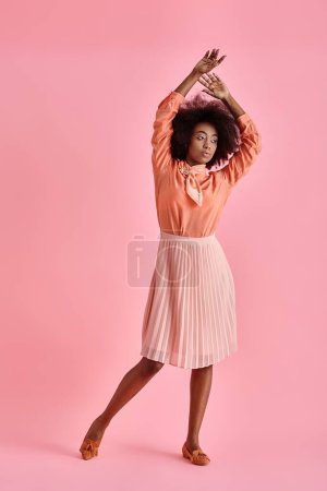 mujer americana africana rizada en blusa de melocotón y falda midi posando con la mano levantada sobre rosa pastel