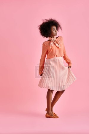 joven afroamericana mujer en blusa de melocotón y falda midi golpea una pose sobre fondo rosa pastel