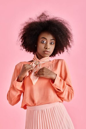 curly african american woman in peach blouse adjusting neck scarf and posing on pink background